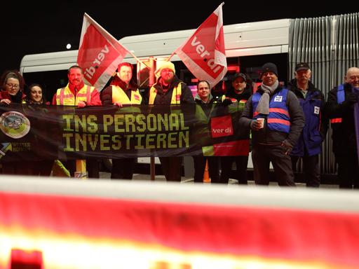 Eine Gruppe Streikender steht in Warnwesten vor einem Bus und hält ein Banner in der Hand, auf dem steht "Ins Personal investieren". 