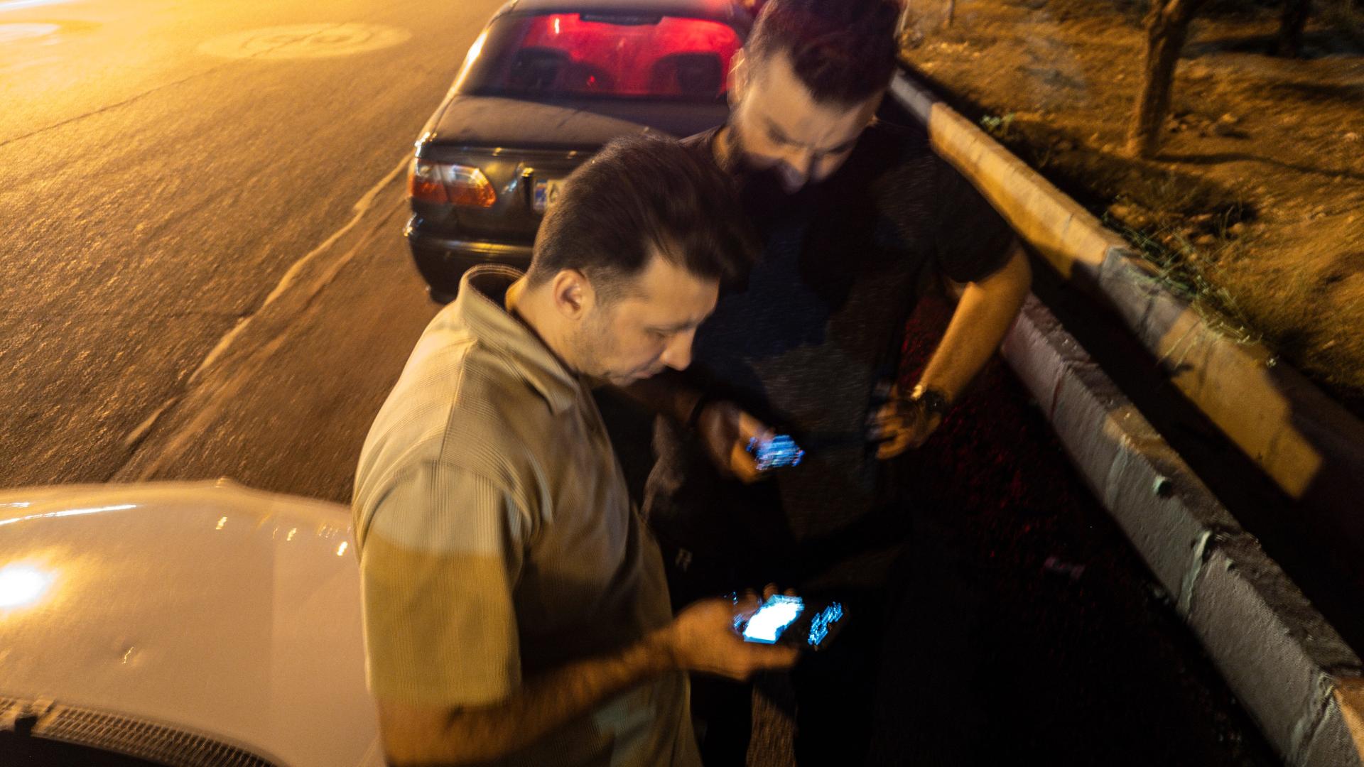 Zwei iranische Männer gucken mitte Juni auf ihre Mobiltelefone, während sie vor einer Tankstelle warten. Im Hintergrund sind die Lichter von Teheran zu erkennen.