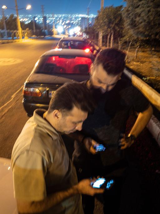Zwei iranische Männer gucken mitte Juni auf ihre Mobiltelefone, während sie vor einer Tankstelle warten. Im Hintergrund sind die Lichter von Teheran zu erkennen.