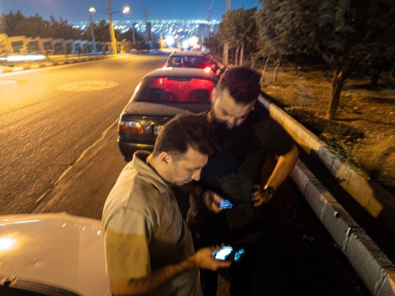 Zwei iranische Männer gucken mitte Juni auf ihre Mobiltelefone, während sie vor einer Tankstelle warten. Im Hintergrund sind die Lichter von Teheran zu erkennen.