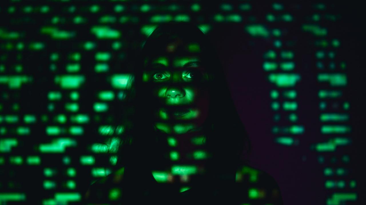 Eine Frau steht an einer Wand in einem dunklen Raum. Auf ihr Gesicht wird mit grünem Licht der Binärcode projiziert.