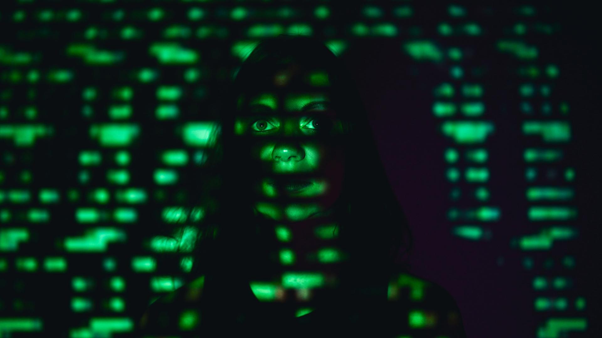 Eine Frau steht an einer Wand in einem dunklen Raum. Auf ihr Gesicht wird mit grünem Licht der Binärcode projiziert. Eine Frau steht an einer Wand in einem dunklen Raum. Auf ihr Gesicht wird mit grünem Licht der Binärcode projiziert.