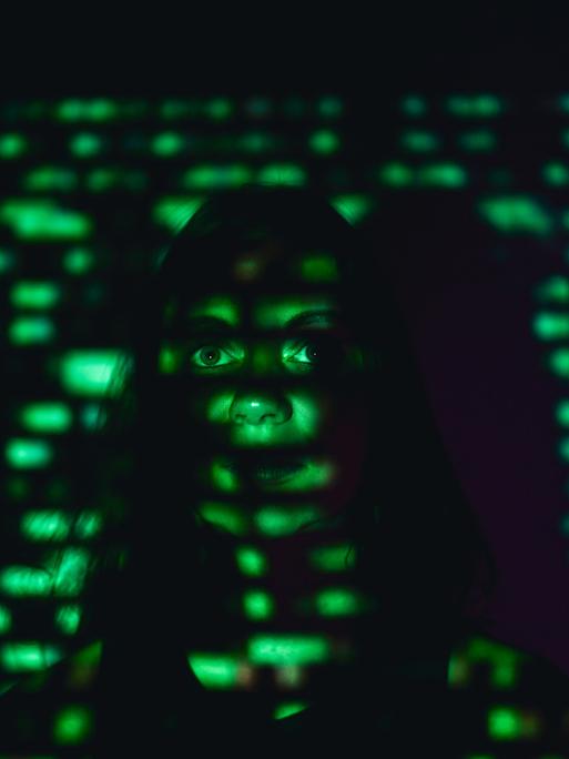Eine Frau steht an eine Wand gelehnt in einem dunklen Raum. Auf ihr Gesicht wird mit grünem Licht der Binärcode projiziert.