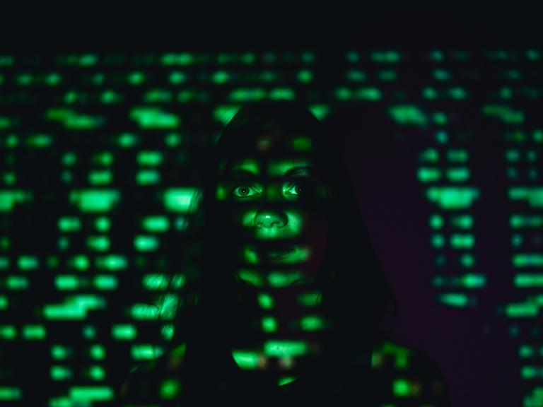 Eine Frau steht an eine Wand gelehnt in einem dunklen Raum. Auf ihr Gesicht wird mit grünem Licht der Binärcode projiziert.