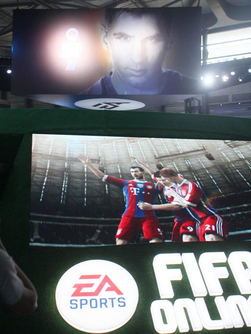 Das Bild zeigt einen Monitor mit virtuellen Spielern des FC Bayern München. Darunter der Name des herstellers. Darüber eine Videowand mit dem gesicht von Lionel Messi. 
