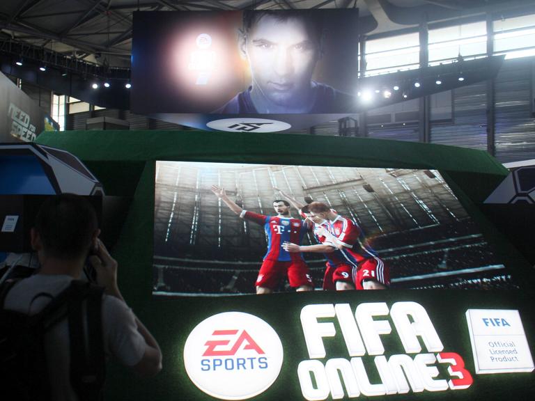 Das Bild zeigt einen Monitor mit virtuellen Spielern des FC Bayern München. Darunter der Name des herstellers. Darüber eine Videowand mit dem gesicht von Lionel Messi. 