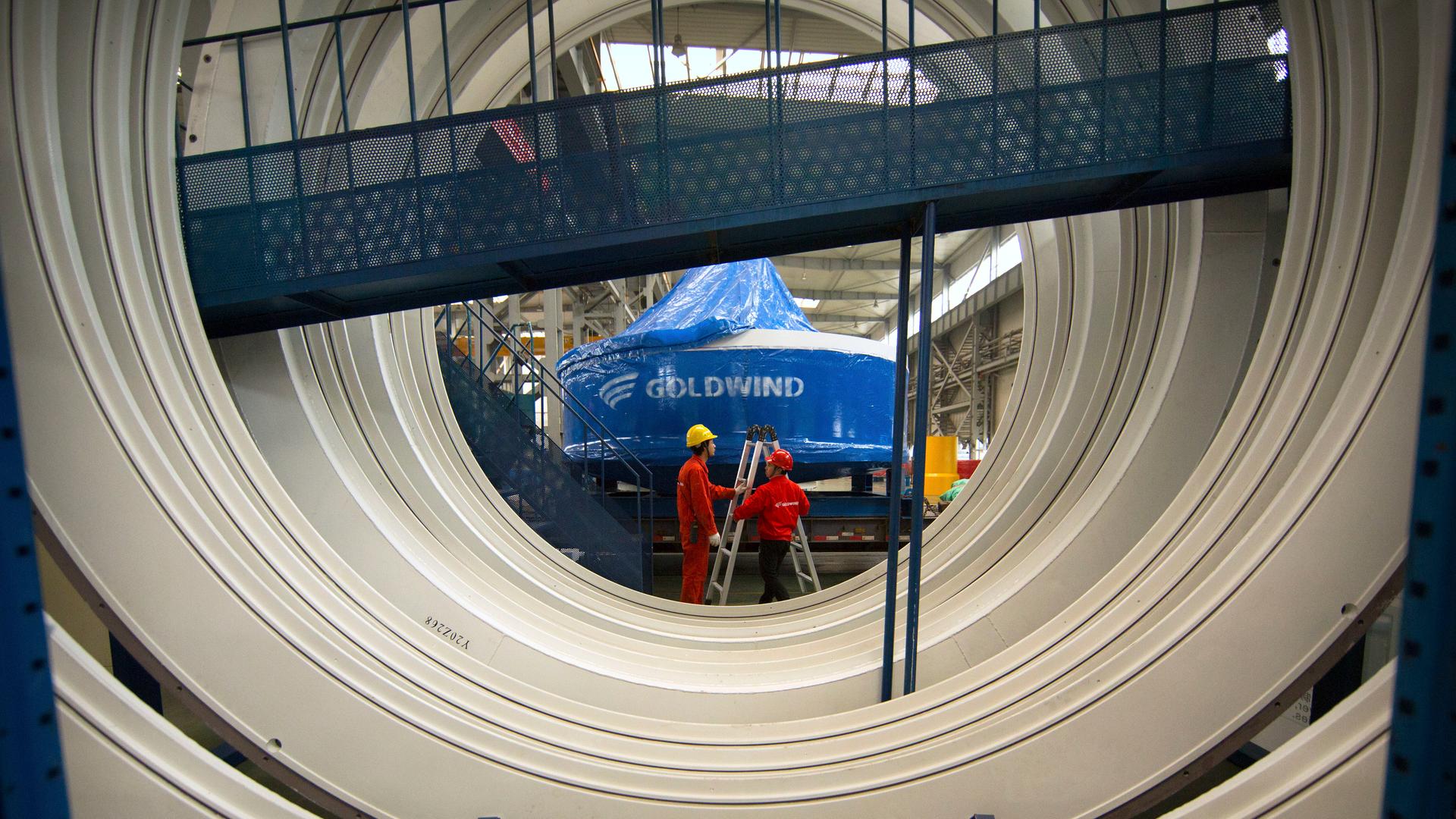 Hinter einer großen Windturbine stehen zwei Arbeiter mit einer Leiter vor der Inschrift "Goldwind".