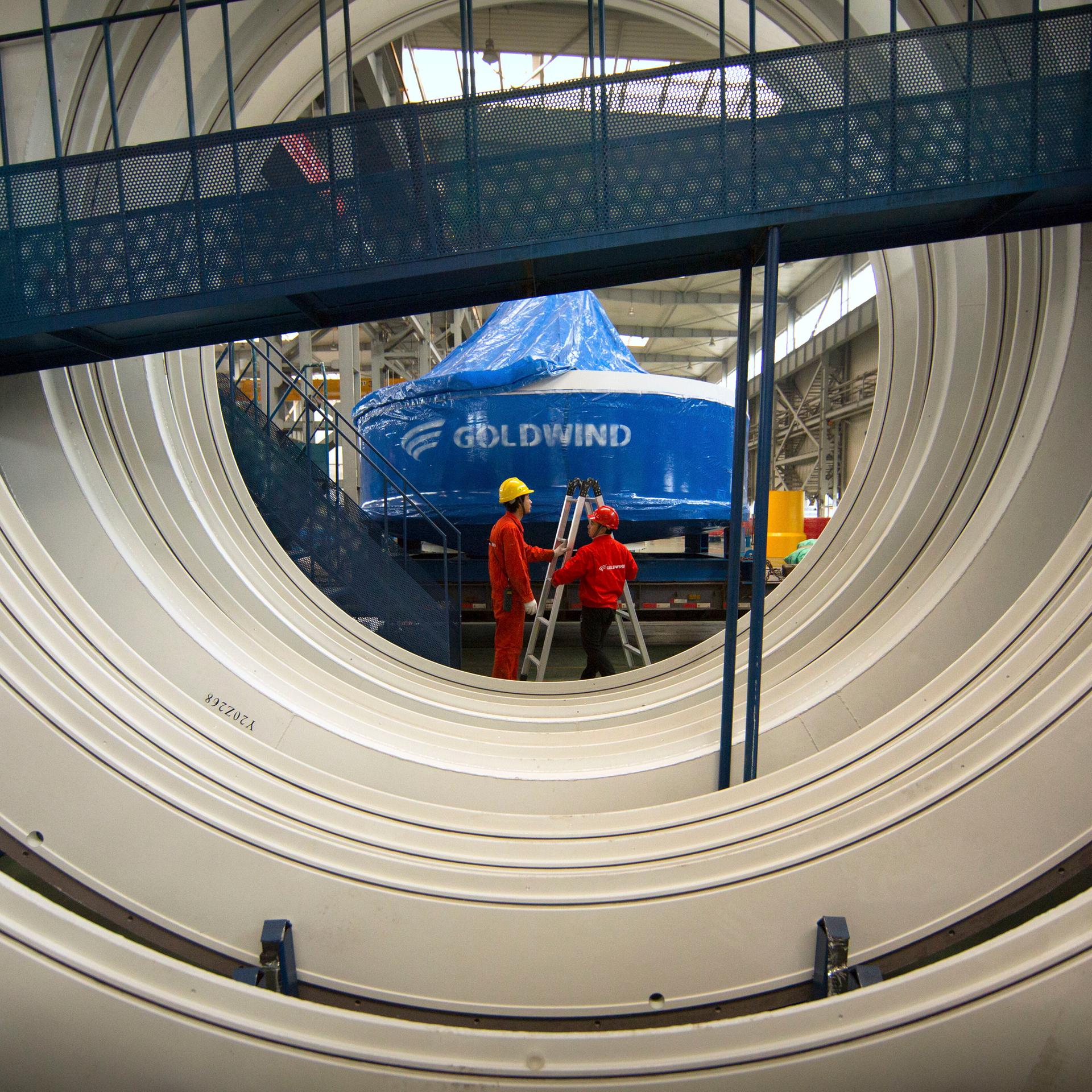 Hinter einer großen Windturbine stehen zwei Arbeiter mit einer Leiter vor der Inschrift "Goldwind".