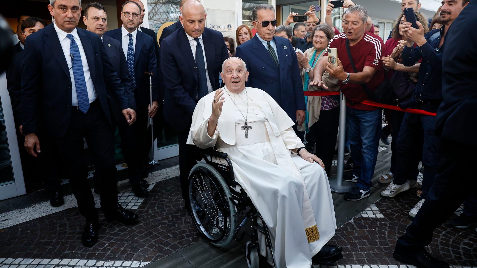 Rom: Papst Franziskus verlässt die Universitätspoliklinik Agostino Gemelli, neun Tage nach einer Bauchoperation. Er sitzt im Rollstuhl und winkt in die Kamera. Um ihn herum steht Sicherheitspersonal und Menschen, die ihn fotografieren. Rom: Papst Franziskus verlässt die Universitätspoliklinik Agostino Gemelli, neun Tage nach einer Bauchoperation. Er sitzt im Rollstuhl und winkt in die Kamera. Um ihn herum steht Sicherheitspersonal und Menschen, die ihn fotografieren.