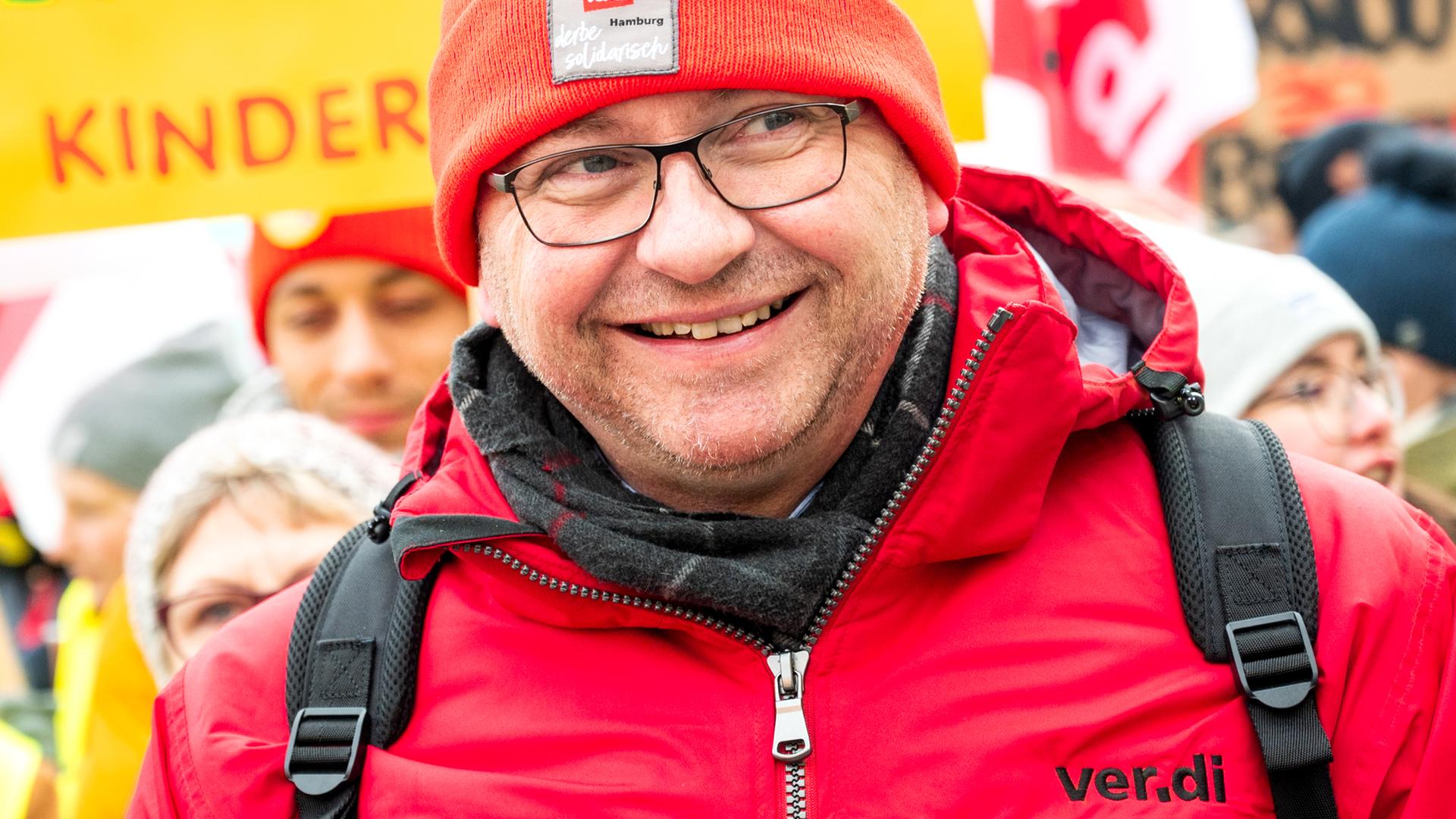 Verdi-Chef Werneke nimmt lächelnd an einer Kundgebung in Schwerin teil, er trägt eine rote Mütze und eine rote Jacke mit der Aufschrift "Verdi". Verdi-Chef Werneke nimmt lächelnd an einer Kundgebung in Schwerin teil, er trägt eine rote Mütze und eine rote Jacke mit der Aufschrift "Verdi".
