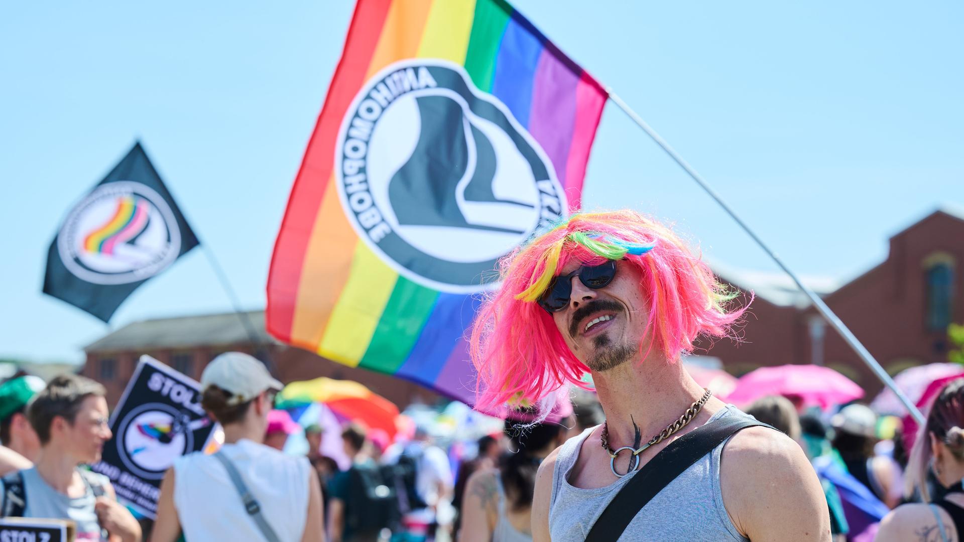 Ein Teilnehmer des Christopher Street Day (CSD) in Eberswalde trägt eine bunte Perücke. Im Hintergrund sieht man eine bunte Flagge. Ein Teilnehmer des Christopher Street Day (CSD) in Eberswalde trägt eine bunte Perücke. Im Hintergrund sieht man eine bunte Flagge.