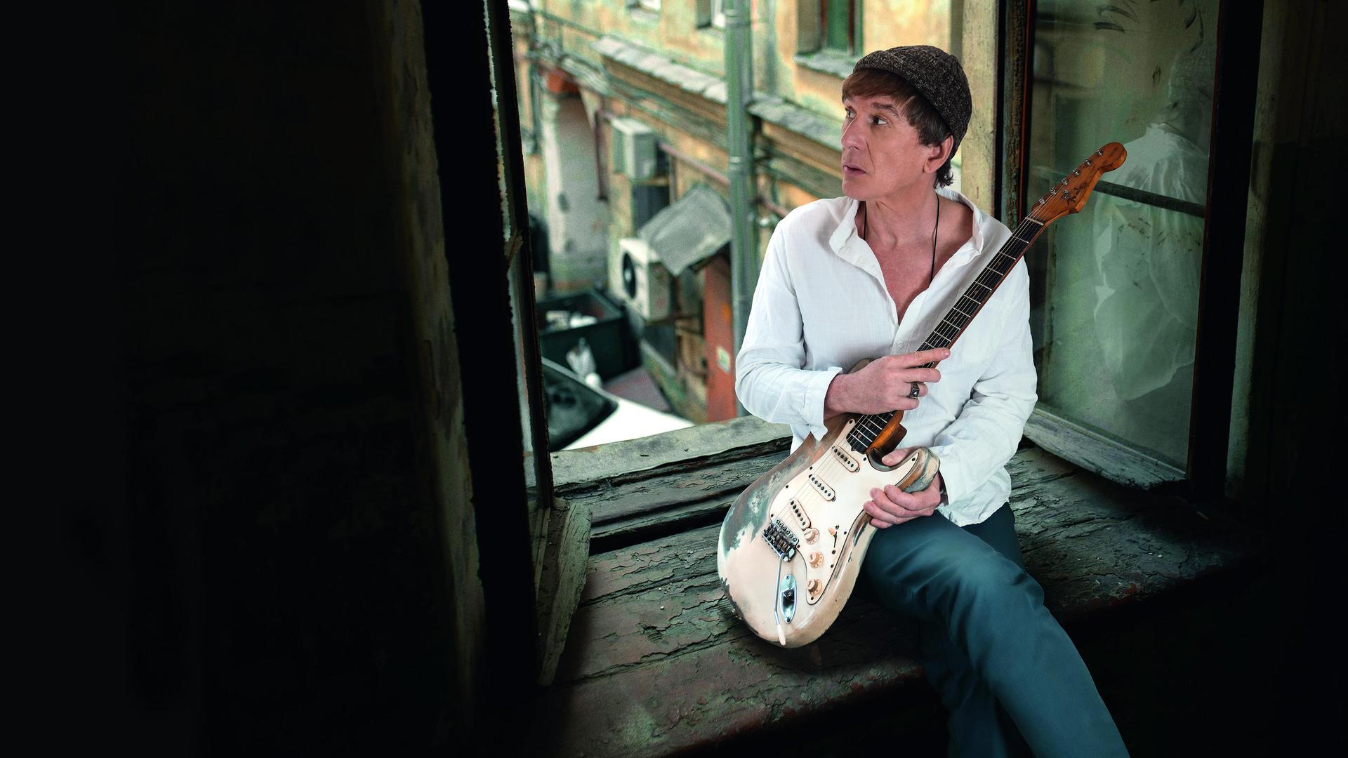 Ein Mann mit E-Gitarre sitzt in einem offenen Fenster. Er trägt ein weißes Hemd, eine Wollmütze über dunkelbraunen Haaren und blickt hinaus in den Innenhof.