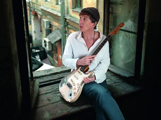 Ein Mann mit E-Gitarre sitzt in einem offenen Fenster. Er trägt ein weißes Hemd, eine Wollmütze über dunkelbraunen Haaren und blickt hinaus in den Innenhof.