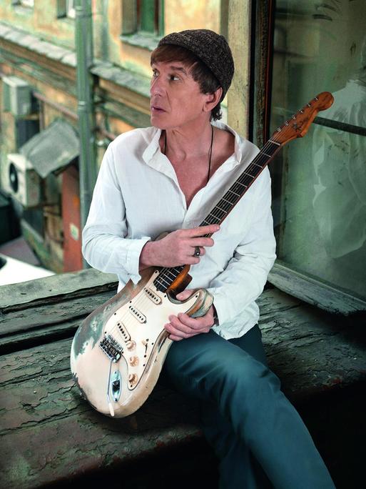Ein Mann mit E-Gitarre sitzt in einem offenen Fenster. Er trägt ein weißes Hemd, eine Wollmütze über dunkelbraunen Haaren und blickt hinaus in den Innenhof.