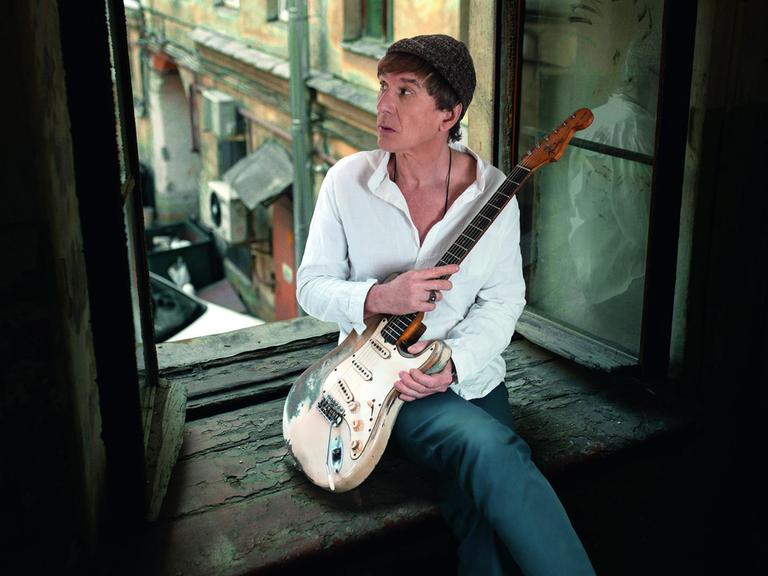 Ein Mann mit E-Gitarre sitzt in einem offenen Fenster. Er trägt ein weißes Hemd, eine Wollmütze über dunkelbraunen Haaren und blickt hinaus in den Innenhof.