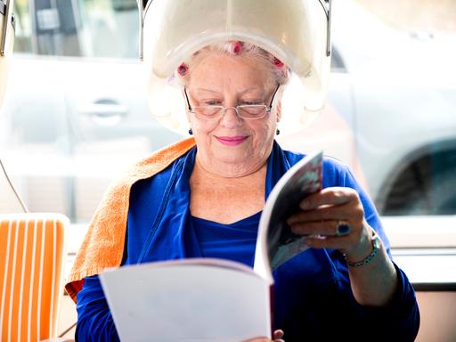Eine ältere Frau sitzt lächelt unter einer Trockenhaube und liest dabei eine Zeitschrift.