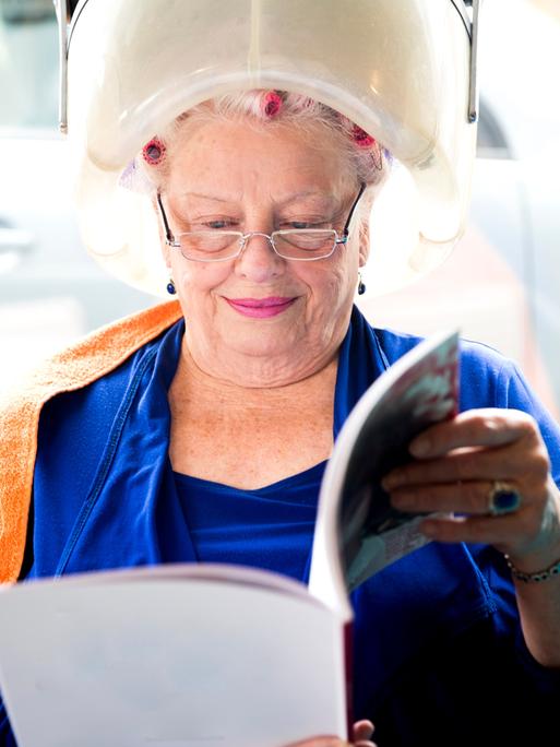 Eine ältere Frau sitzt lächelt unter einer Trockenhaube und liest dabei eine Zeitschrift.