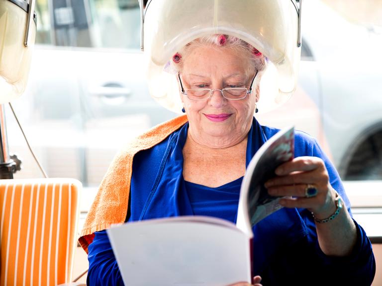 Eine ältere Frau sitzt lächelt unter einer Trockenhaube und liest dabei eine Zeitschrift.