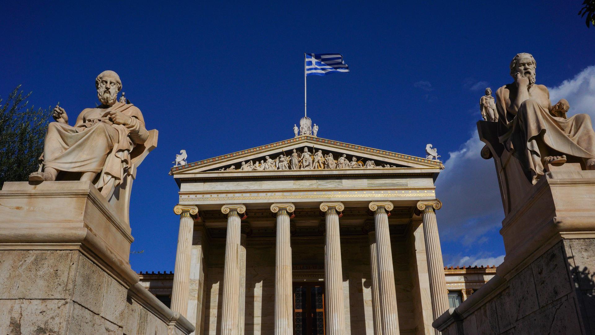 Die Statuen von Sokrates und Platon im Hof der Akademie in Athen vor blauem Himmel.