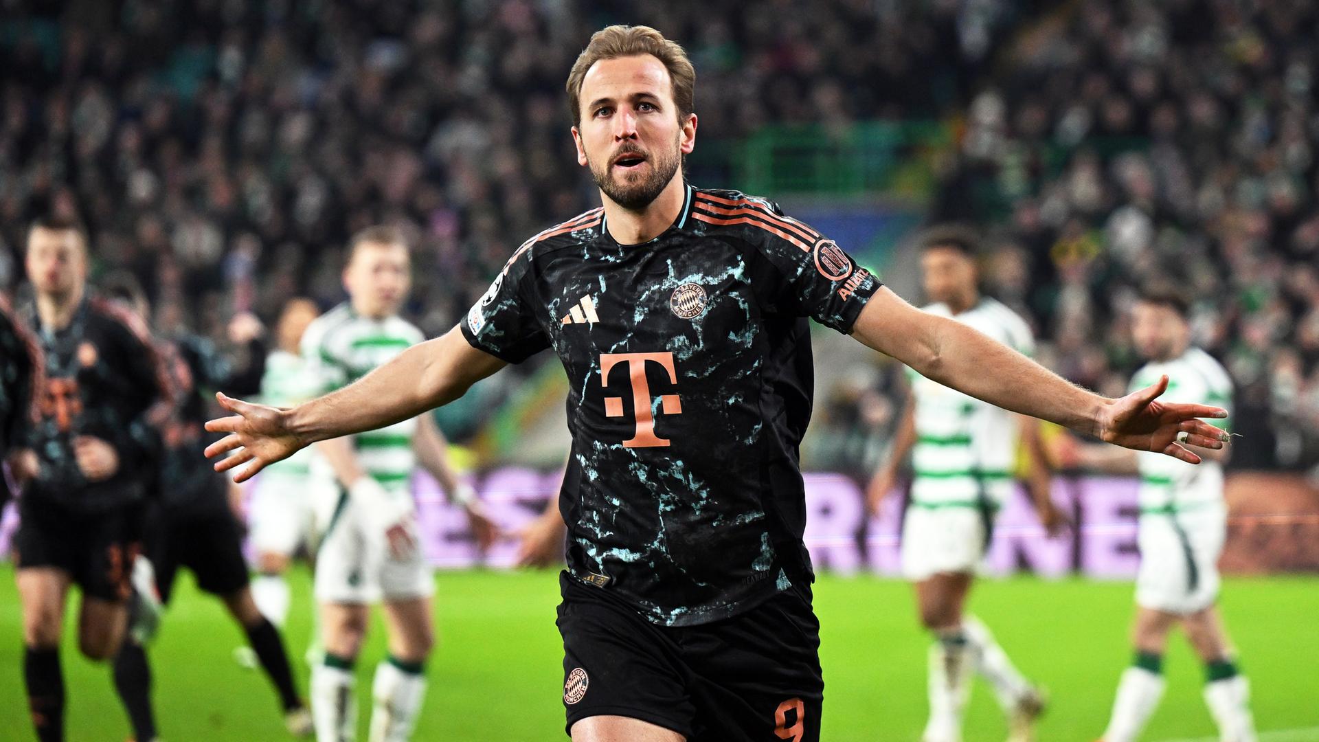 Kane im schwarz-roten Trikot breitet beide Arme aus und läuft auf den Betrachter zu. Dahinter unscharf Spieler beider Manschaften und eine vollbesetzte Tribüne.