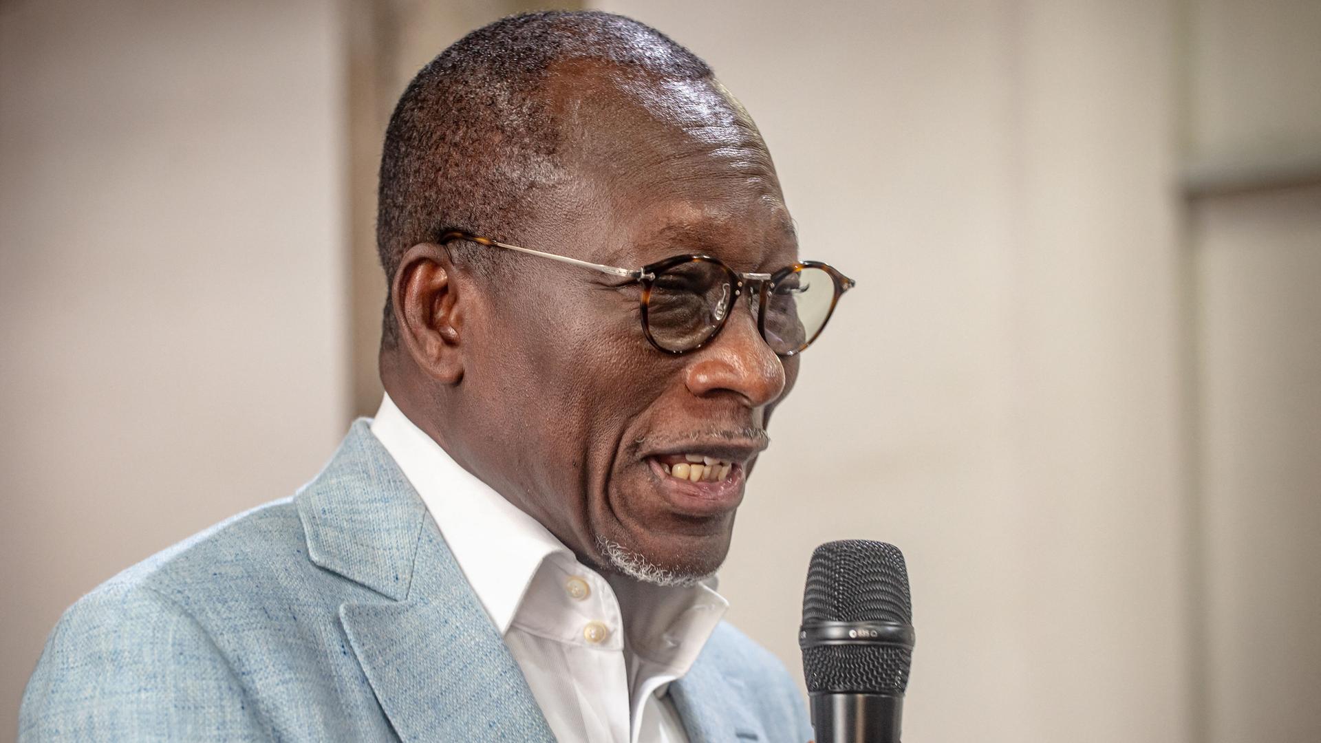 Ein schwarzer Mann mit kurzen Haaren und runder Brille spricht in ein Mikrofon. Er trägt ein weißes Hemd und ein graues Sakko. Ein schwarzer Mann mit kurzen Haaren und runder Brille spricht in ein Mikrofon. Er trägt ein weißes Hemd und ein graues Sakko.