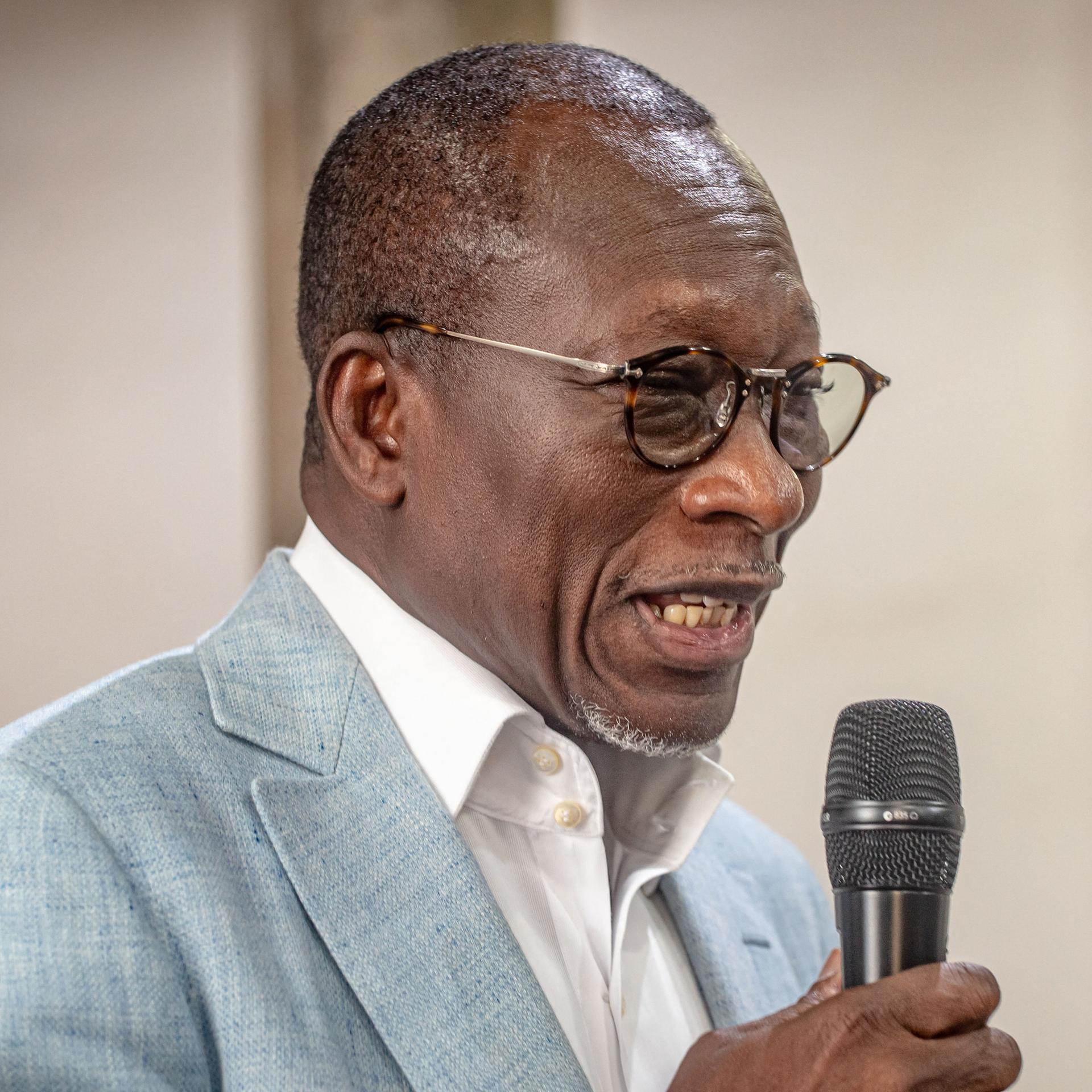 Ein schwarzer Mann mit kurzen Haaren und runder Brille spricht in ein Mikrofon. Er trägt ein weißes Hemd und ein graues Sakko.