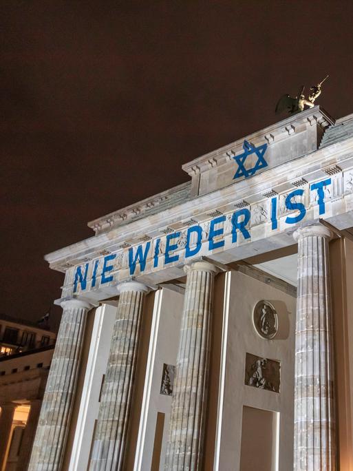 Das Brandenburger Tor ist blau-weiß angestrahlt. Der Schriftzug "Nie wieder ist Jetzt" ist zu lesen. 