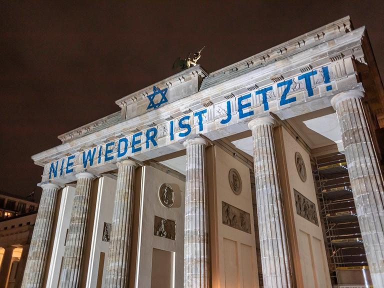 Das Brandenburger Tor ist blau-weiß angestrahlt. Der Schriftzug "Nie wieder ist Jetzt" ist zu lesen. 