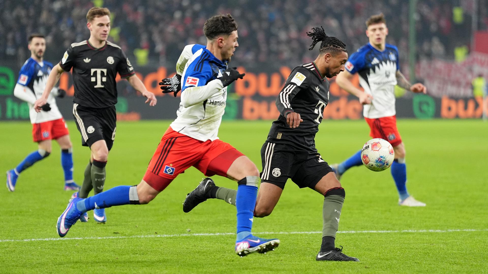 Das Bild zeigt zwei Fußball-Spieler in vollem Lauf beim Kampf um den Ball. In Hintergrund sind Mitspieler zu sehen, die das Geschehen beobachten.