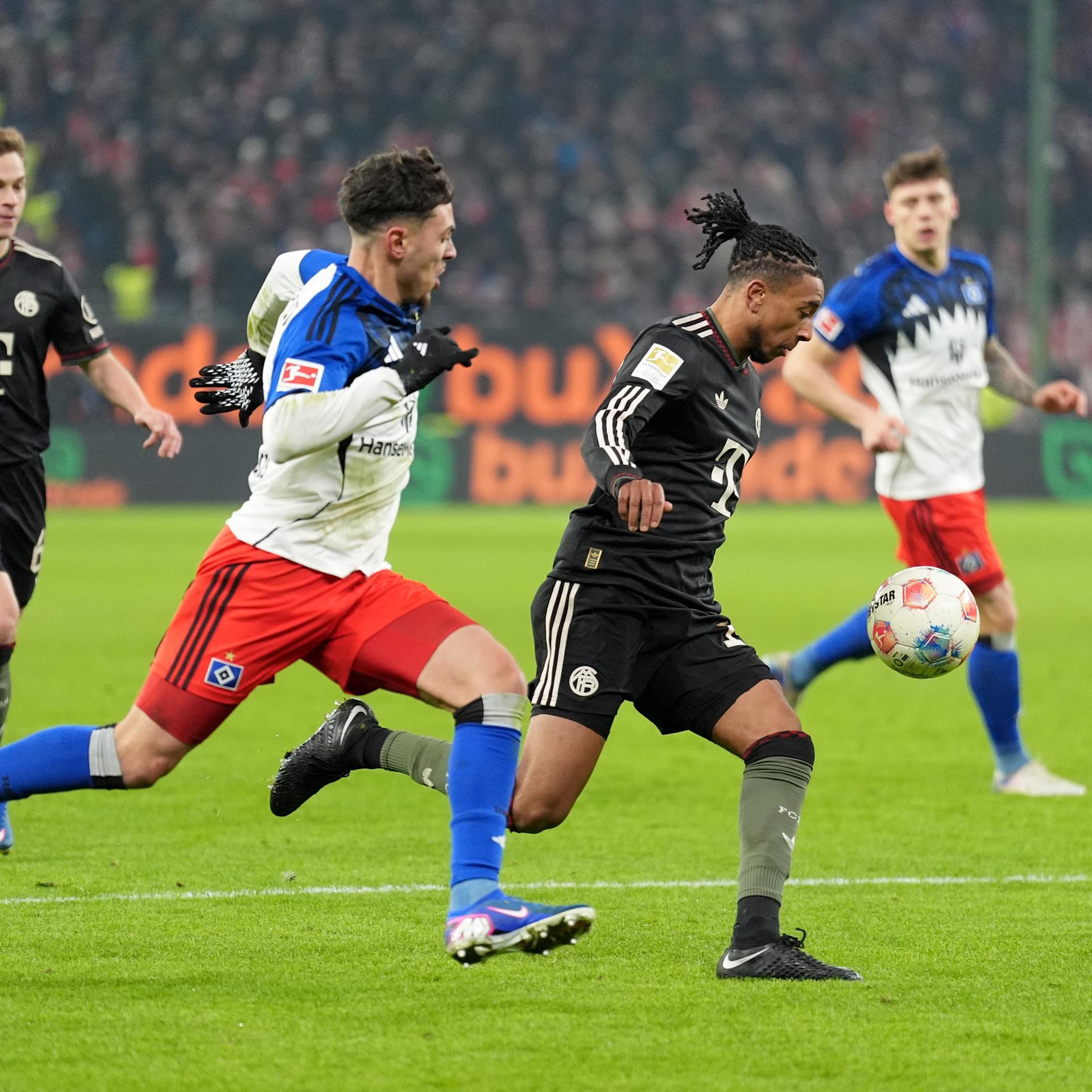 Das Bild zeigt zwei Fußball-Spieler in vollem Lauf beim Kampf um den Ball. In Hintergrund sind Mitspieler zu sehen, die das Geschehen beobachten.