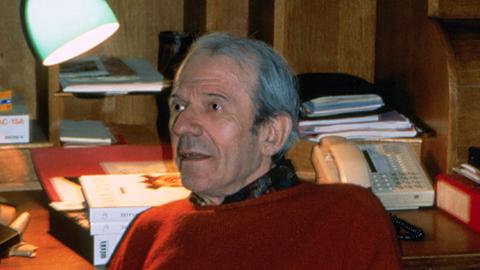 Der Philosoph Gilles Deleuze sitzt in einem rotem Pullover in Paris vor einem Schreibtisch. Im Hintergrund ist eine Lampe und ein Telefon.