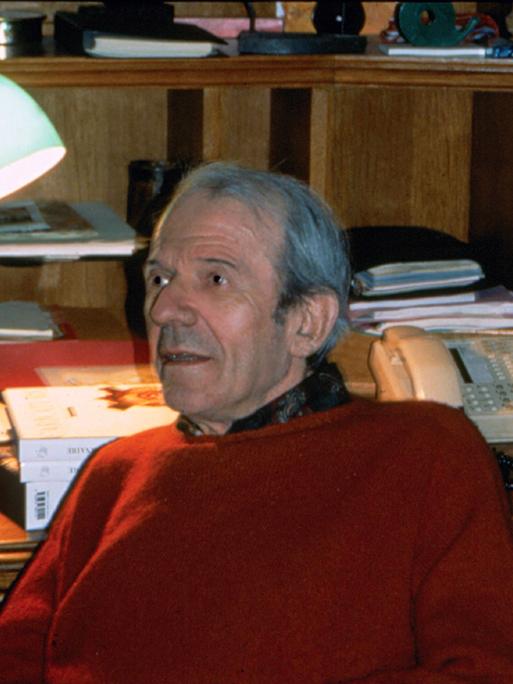 Der Philosoph Gilles Deleuze sitzt in einem rotem Pullover in Paris vor einem Schreibtisch. Im Hintergrund ist eine Lampe und ein Telefon.