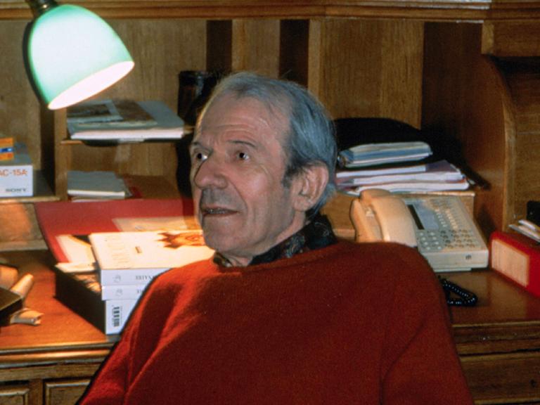 Der Philosoph Gilles Deleuze sitzt in einem rotem Pullover in Paris vor einem Schreibtisch. Im Hintergrund ist eine Lampe und ein Telefon.