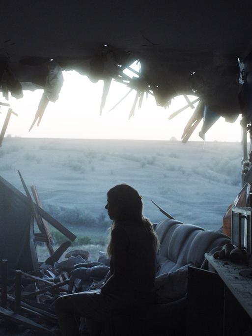 Eine junge Frau sitzt in einem zerstörtem Wohnzimmer und schaut durch eine zerstörte Hauswand nach draußen.
