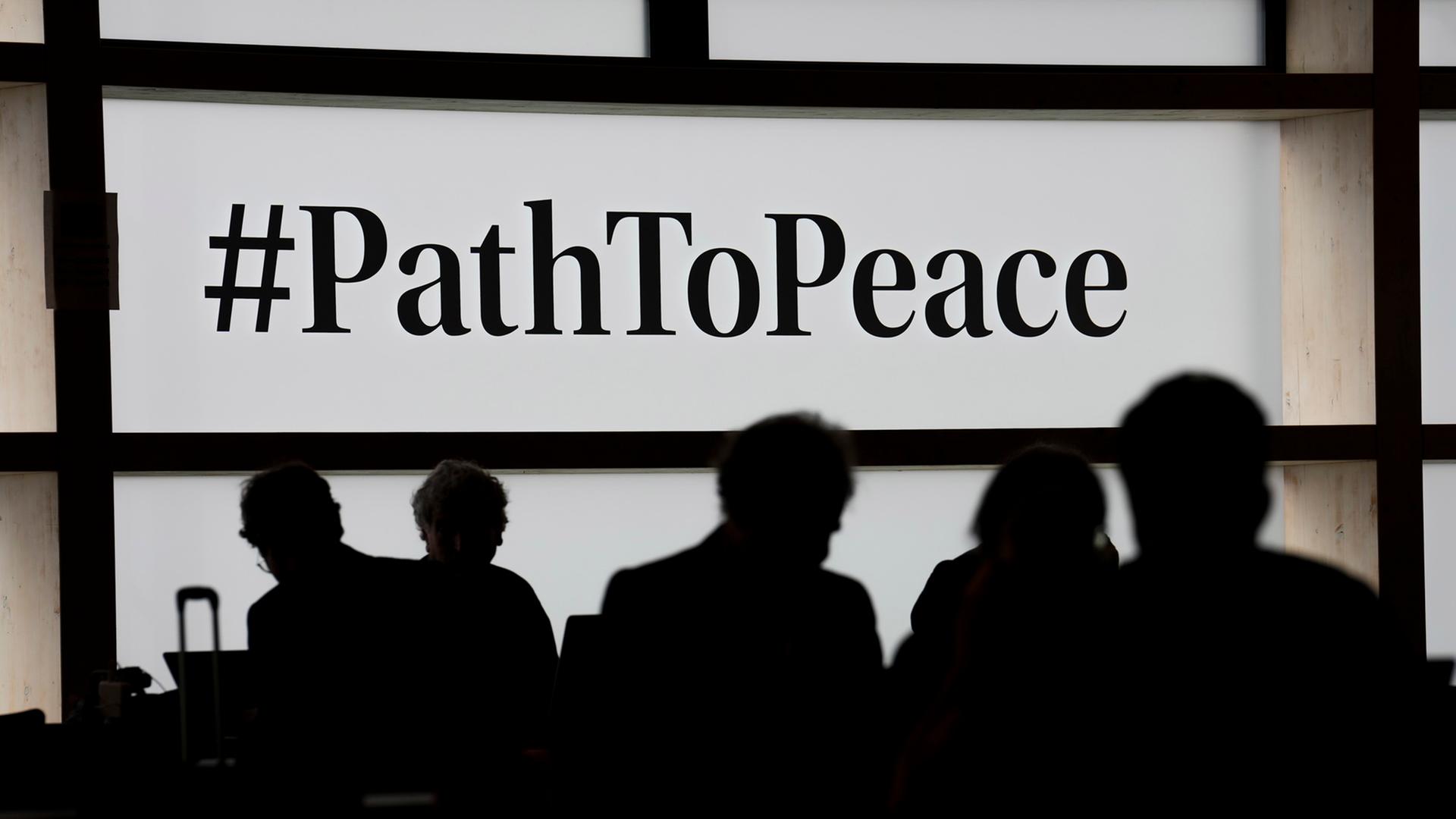 Das Bild zeigt die Silhouette mehrerer Menschen vor einer Leindwand mit der Aufschrift "#pathtopeace".