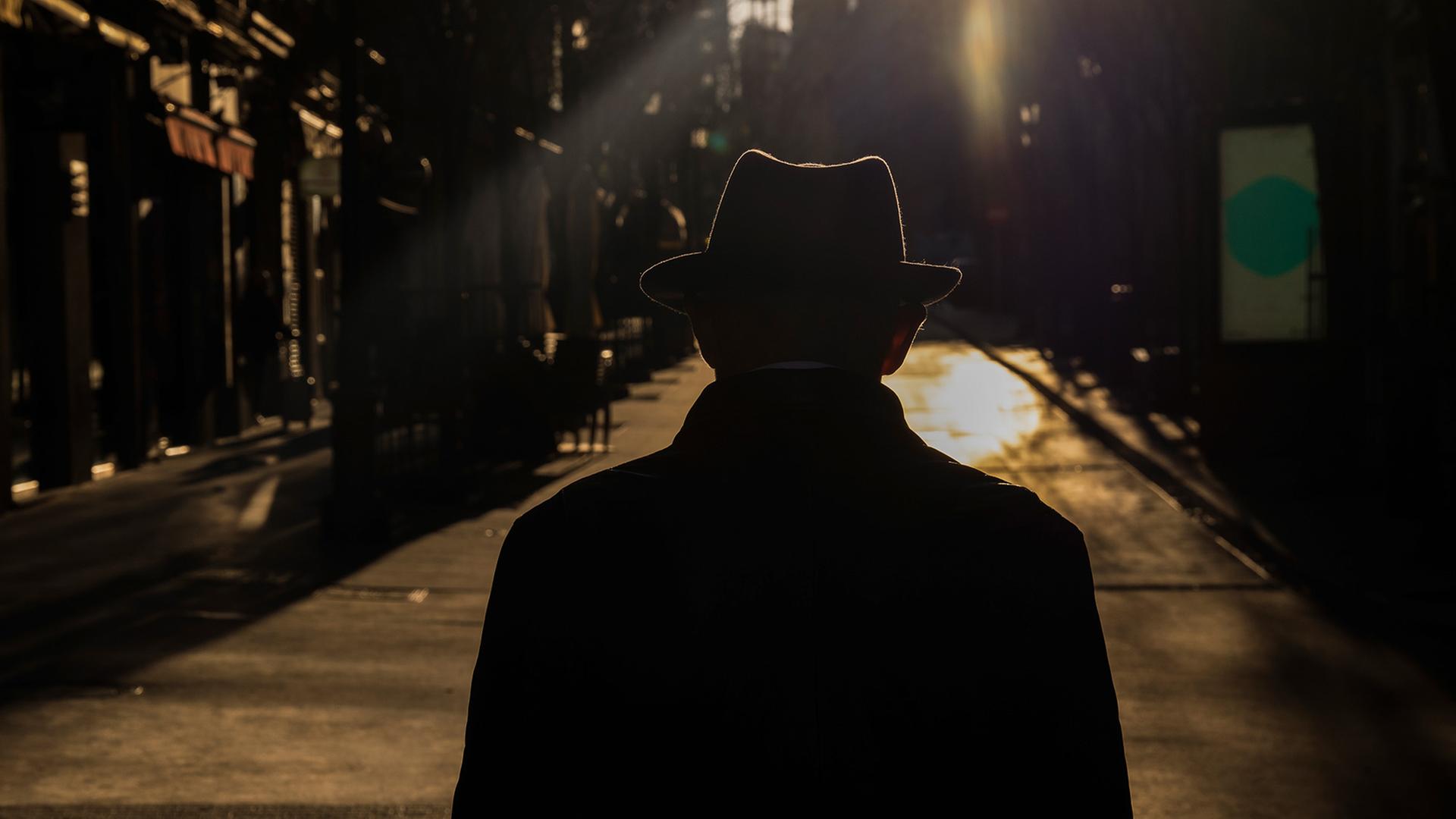 Ein Mann mit Hut geht mit dem Rücken zur Kamera eine Straße in der Dunkelheit entlang. 