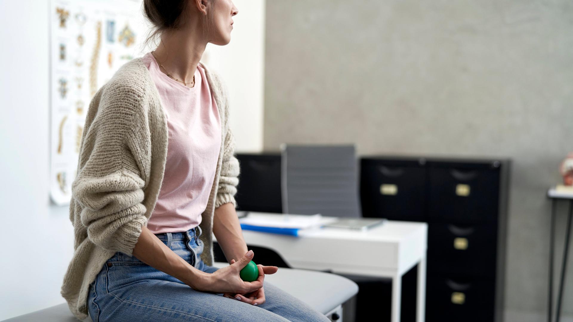Eine Frau sitzt in einem Behandlungszimmer beim Arzt auf einer Liege. Sie wartet dort und hält dabei einen kleinen grünen Gummiball in den Händen.