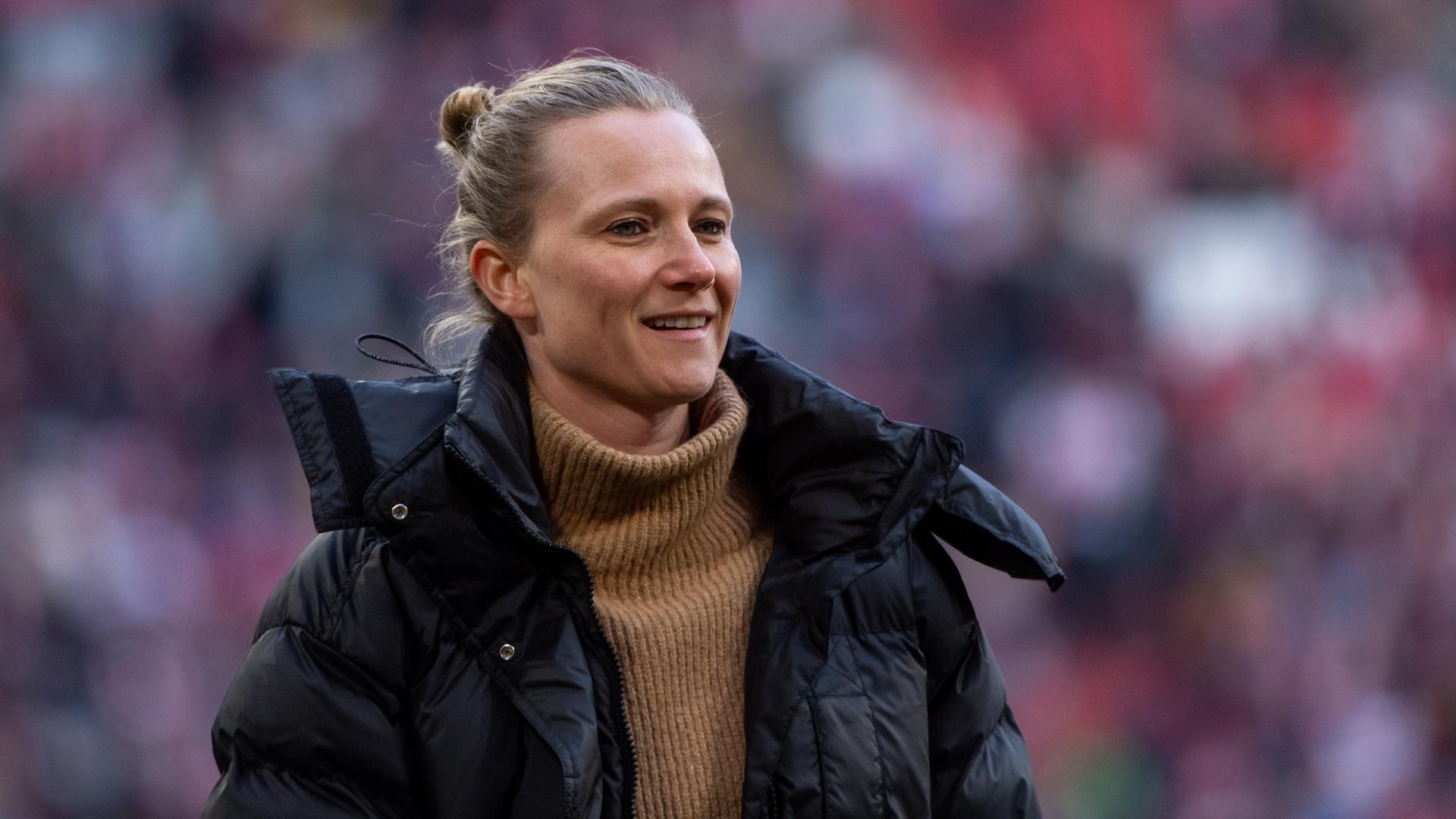 Kathleen Krüger steht am Spielfeldrand, hinter ihr die Tribüne mit FC Bayern-Fans