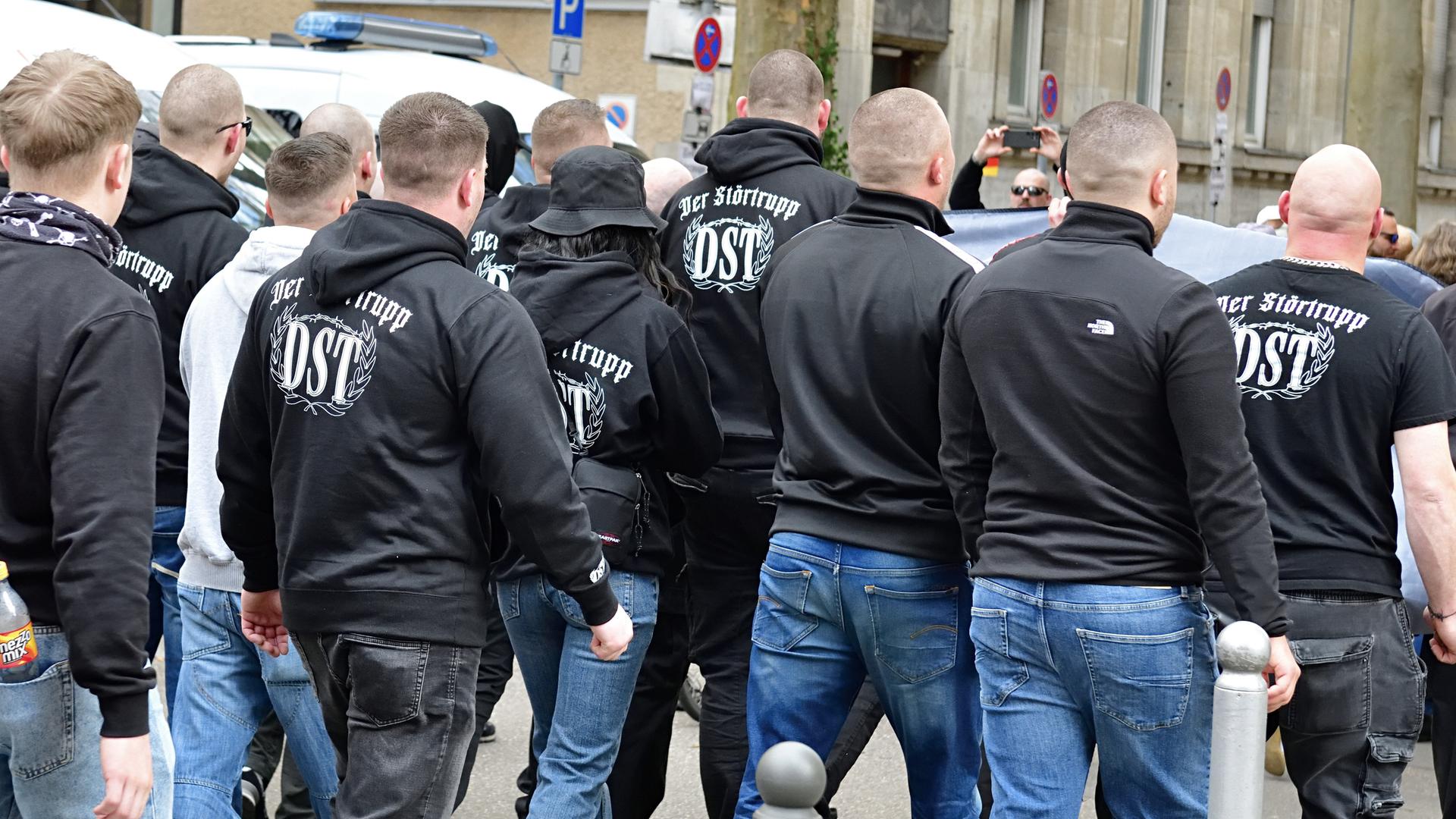 Das Bild zeigt circa 13 Personen, die auf einer Demo marschieren, von hinten. Die meisten tragen dunkle Kapuzenpollover mit der Rückenaufschrift „Der Störtrupp – DST“ Das Bild zeigt circa 13 Personen, die auf einer Demo marschieren, von hinten. Die meisten tragen dunkle Kapuzenpollover mit der Rückenaufschrift „Der Störtrupp – DST“