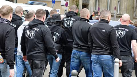 Das Bild zeigt circa 13 Personen, die auf einer Demo marschieren, von hinten. Die meisten tragen dunkle Kapuzenpollover mit der Rückenaufschrift „Der Störtrupp – DST“