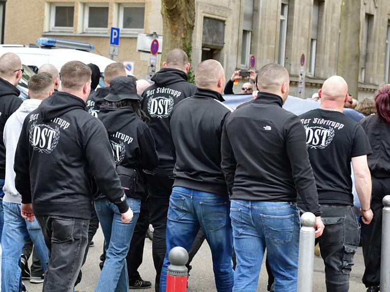 Das Bild zeigt circa 13 Personen, die auf einer Demo marschieren, von hinten. Die meisten tragen dunkle Kapuzenpollover mit der Rückenaufschrift „Der Störtrupp – DST“
