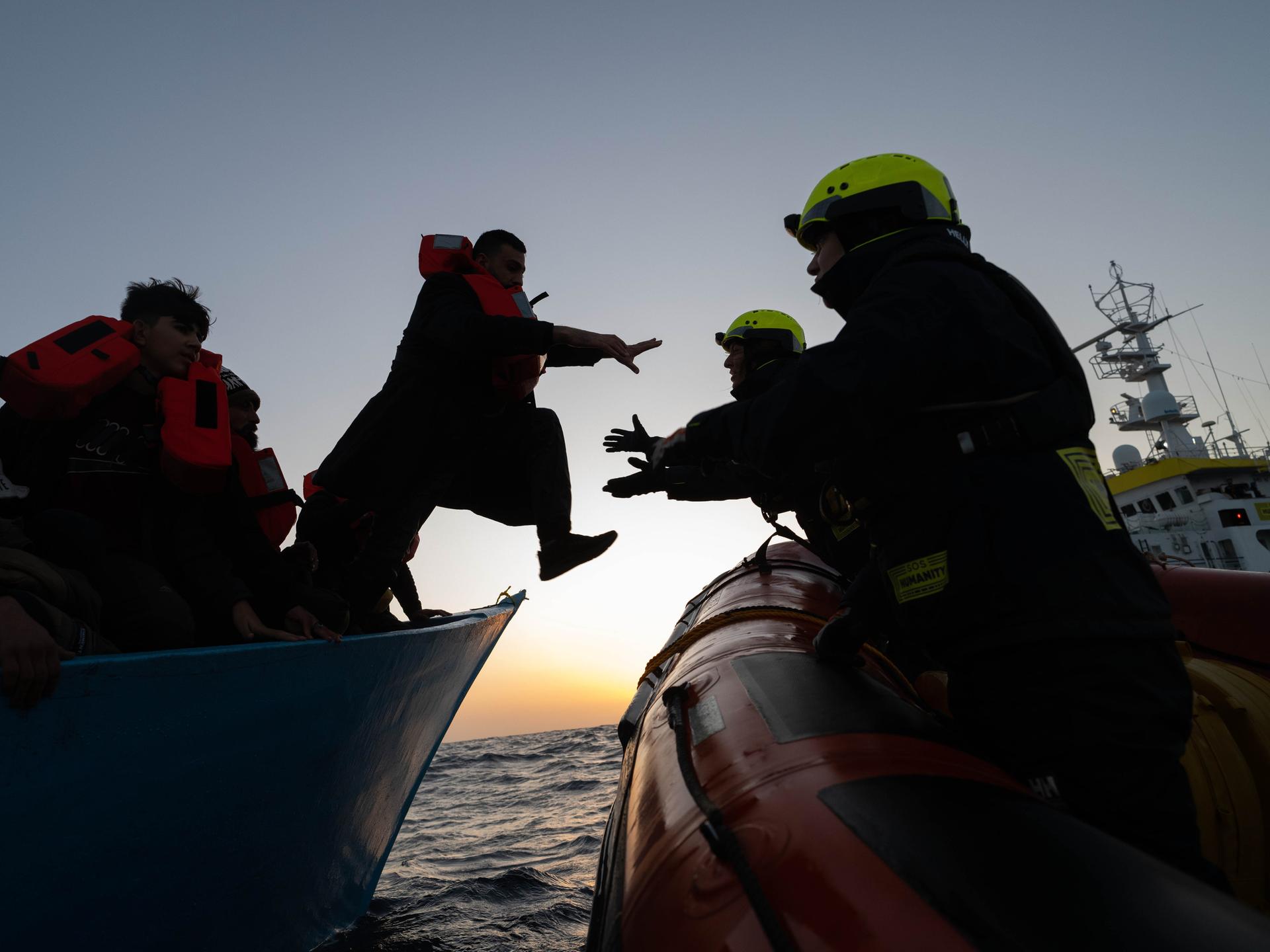 Zwei Personen mit Schutzhelmen auf einem Boot nehmen eine Person mit Rettungsweste aus einem anderen Boot in Empfang. Es sitzen weitere Personen mit Rettungswesten im Boot. Die Personen sind nur als Silhouetten vor dem Abendhimmel auf dem Meer umrissen.
