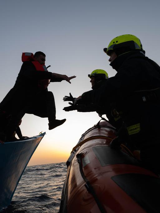 Zwei Personen mit Schutzhelmen auf einem Boot nehmen eine Person mit Rettungsweste aus einem anderen Boot in Empfang. Es sitzen weitere Personen mit Rettungswesten im Boot. Die Personen sind nur als Silhouetten vor dem Abendhimmel auf dem Meer umrissen.