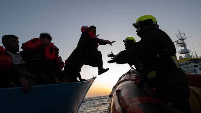 Zwei Personen mit Schutzhelmen auf einem Boot nehmen eine Person mit Rettungsweste aus einem anderen Boot in Empfang. Es sitzen weitere Personen mit Rettungswesten im Boot. Die Personen sind nur als Silhouetten vor dem Abendhimmel auf dem Meer umrissen.