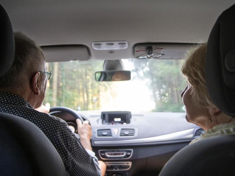 Ein Rentner-Ehepaar sitzt im Auto. Der Mann fährt, die Frau schaut zu ihm herüber.