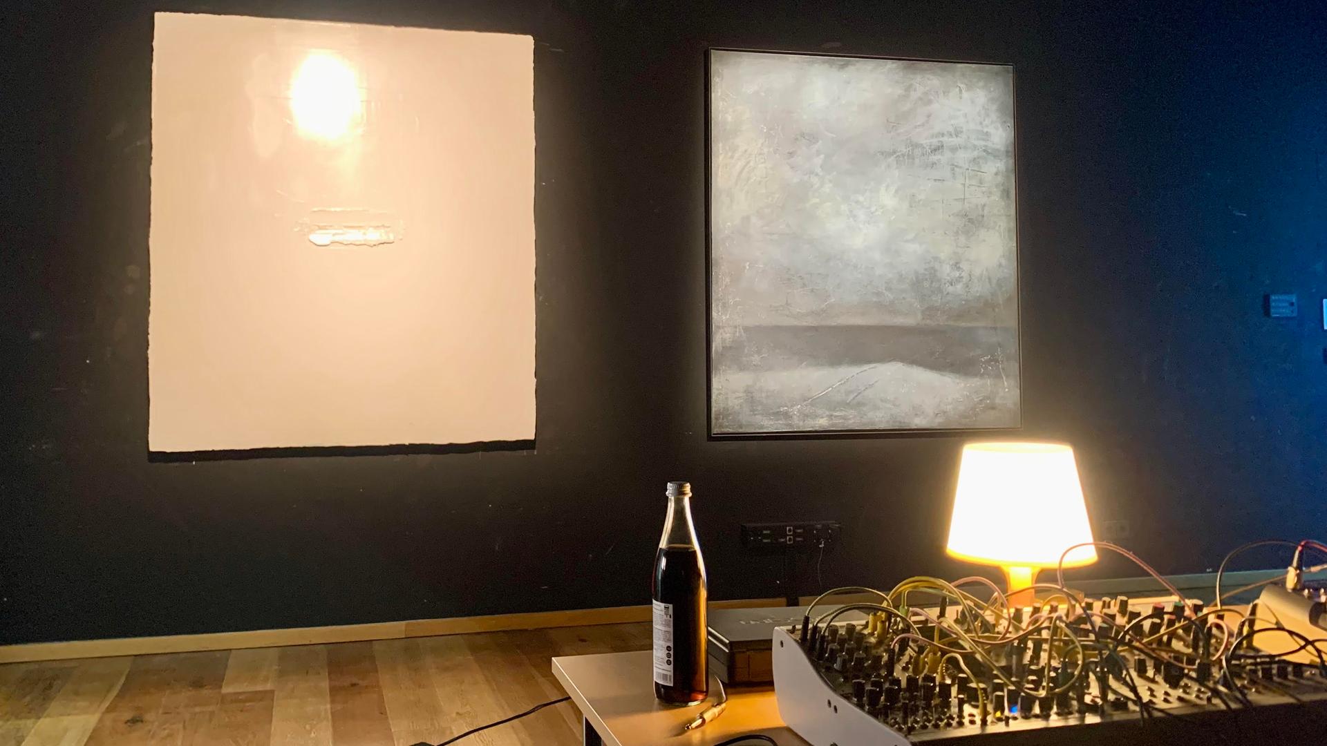Ein dunkler Ausstellungsraum, an der Wand zwei illuminierte Bilder, im Vordergrund ein Mischpult, daneben eine Bierflasche