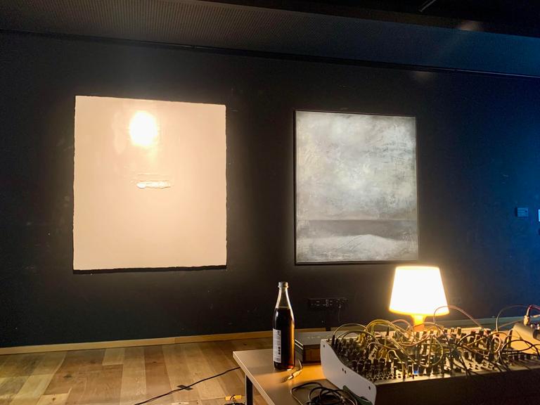Ein dunkler Ausstellungsraum, an der Wand zwei illuminierte Bilder, im Vordergrund ein Mischpult, daneben eine Bierflasche