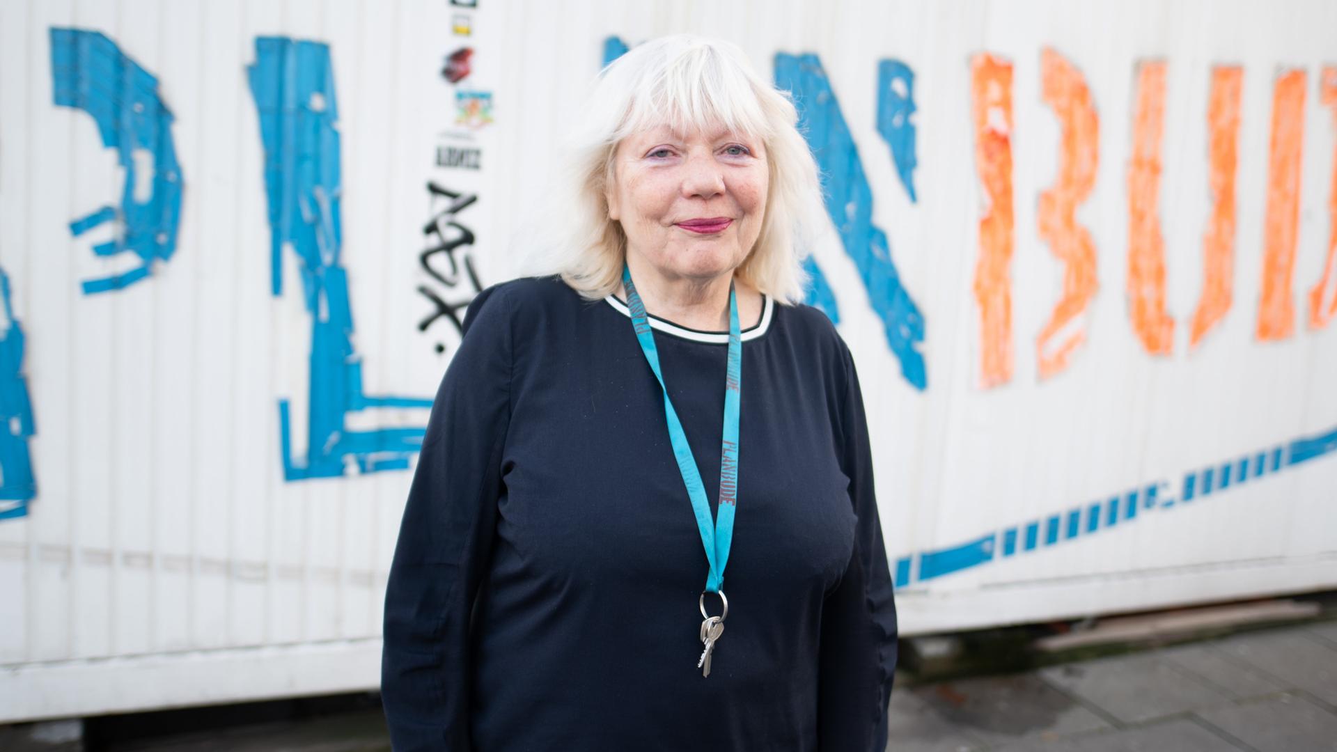 Die Künstlerin Margit Czenki steht vor einem weißen Container mit der Aufschrift "Planbude". Um den Hals trägt sie eine Kette mit einem Schlüssel.