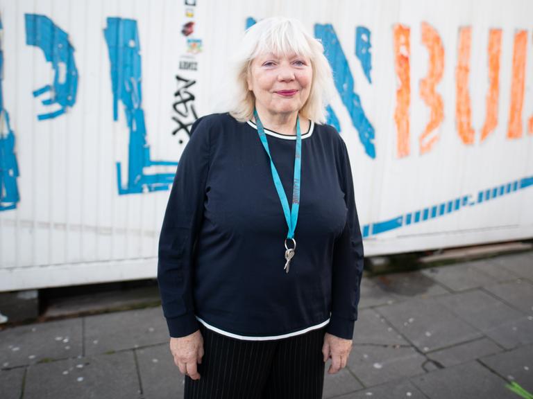 Die Künstlerin Margit Czenki steht vor einem weißen Container mit der Aufschrift "Planbude". Um den Hals trägt sie eine Kette mit einem Schlüssel.