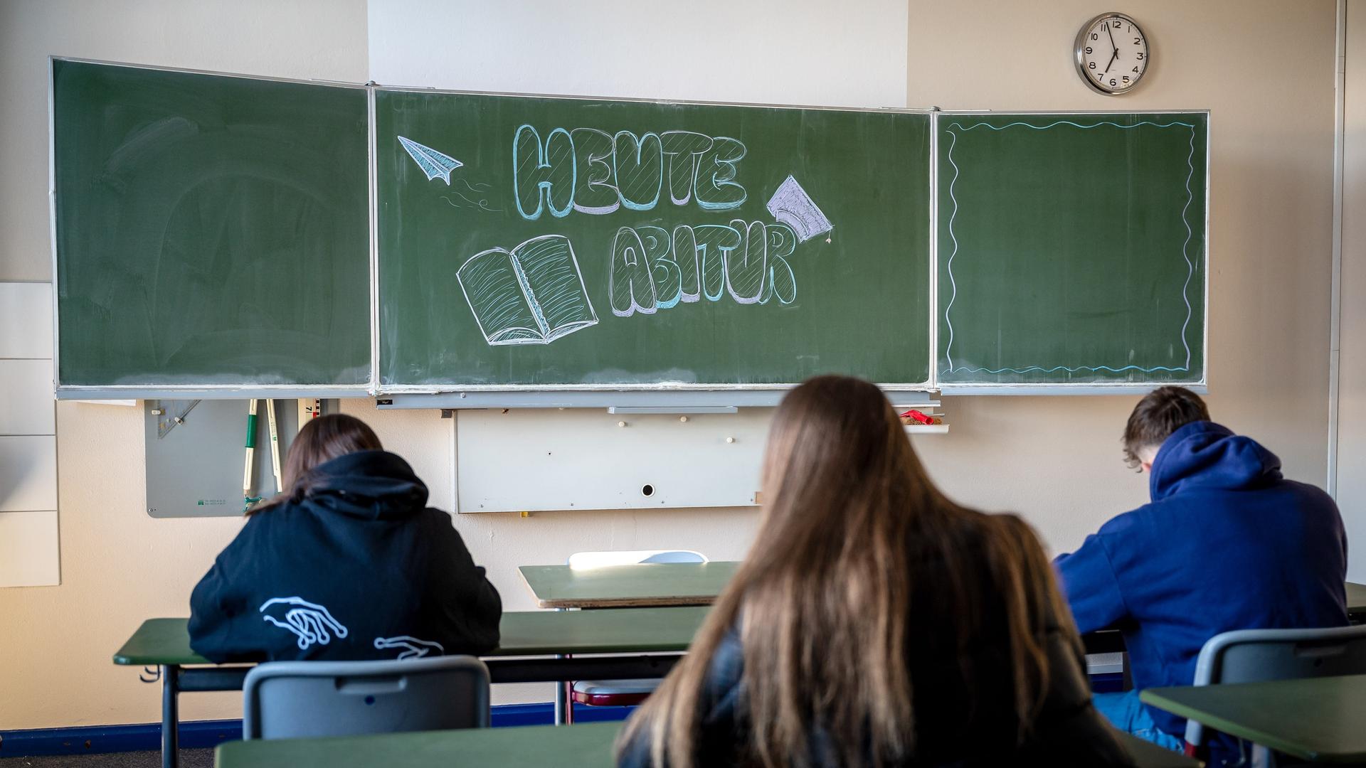 "Heute Abitur" steht auf einer Tafel im Klassenzimmer eines Gymnasiums in Niedersachsen. Vor der Tafel sitzen drei Schüler an ihren Schreibtischen. (Symbolbild)